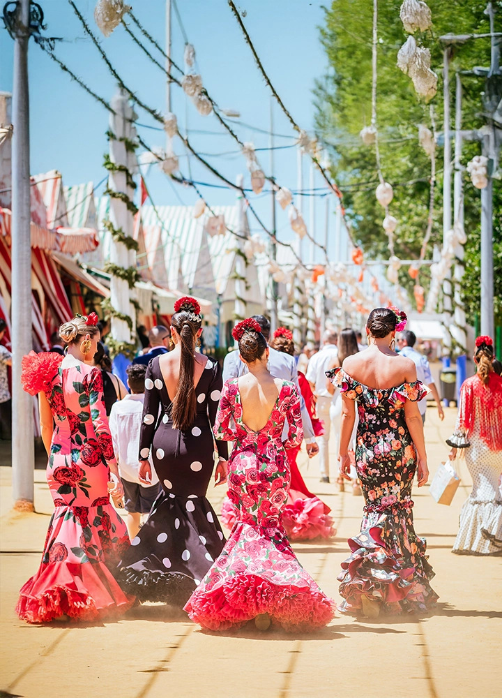 Cuatro mujeres vestidas con trajes de flamenca caminan por una calle decorada con guirnaldas y farolillos durante una feria. Los trajes son coloridos, con estampados florales y lunares, y tienen volantes en las mangas y la falda. La calle está llena de casetas típicas de feria al fondo.