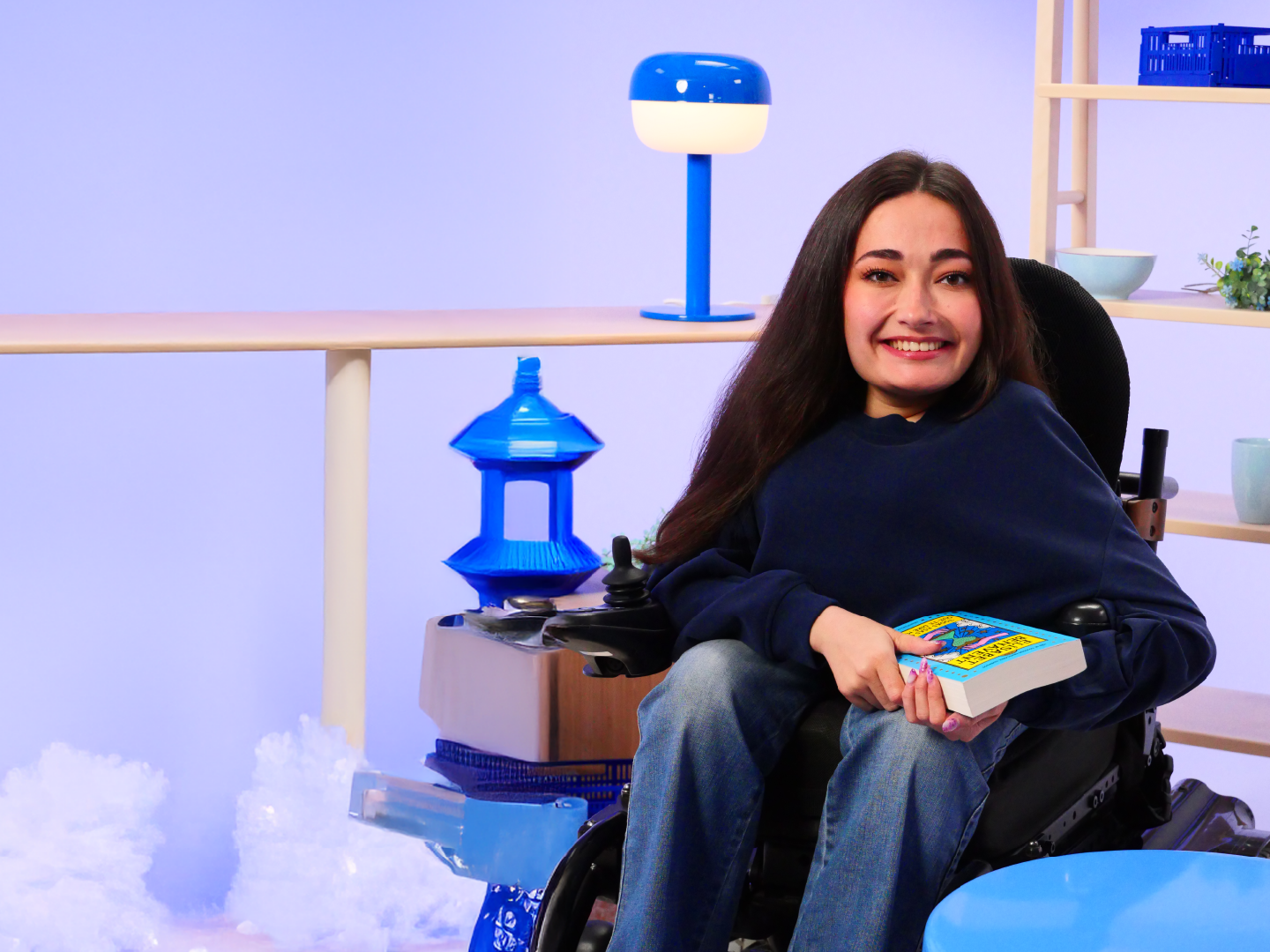 persona sentada en una silla de ruedas, sosteniendo un libro con una portada colorida que tiene texto impreso. El fondo incluye varios objetos cuidadosamente dispuestos, como una lámpara azul, un adorno azul que recuerda a una pagoda, y estanterías con tazones y plantas. El entorno tiene una estética organizada con un predominante tono azul claro, lo que le da una sensación tranquila e interior.
