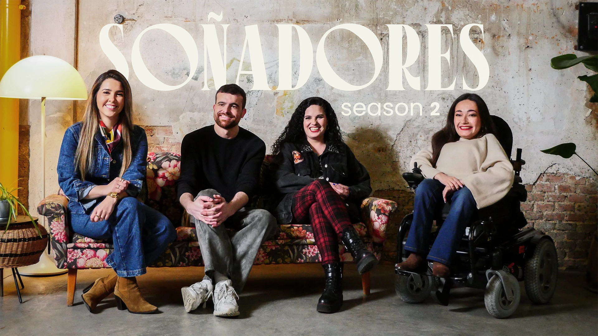Cuatro personas sonrientes están sentadas en un sofá vintage con estampado floral frente a una pared de cemento con textura envejecida. Sobre ellos, en grandes letras blancas, se lee “SOÑADORES season 2”. A la izquierda, una mujer con chaqueta vaquera, pañuelo de colores y botas marrones. A su lado, un hombre con jersey negro y pantalones grises, con las manos entrelazadas. Junto a él, una mujer de cabello rizado y chaqueta negra con tachuelas, vistiendo pantalón de cuadros y botas negras. A la derecha, una mujer con blusa beige y pantalón azul, sentada en una silla de ruedas eléctrica, con una gran sonrisa. La imagen refleja un ambiente acogedor y auténtico, con un estilo industrial y bohemio.
