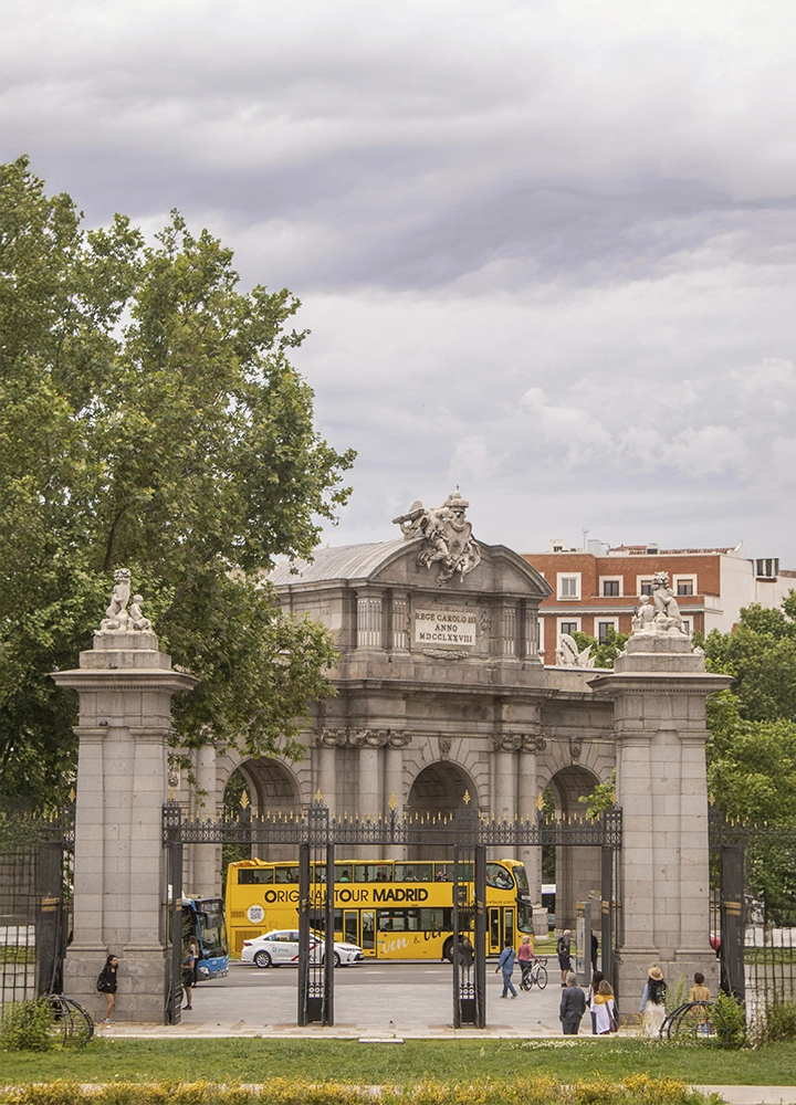 Vista de la Puerta de Alcalá, un monumento de piedra con arcos y esculturas, con autobuses turísticos amarillos pasando y gente caminando por la zona.