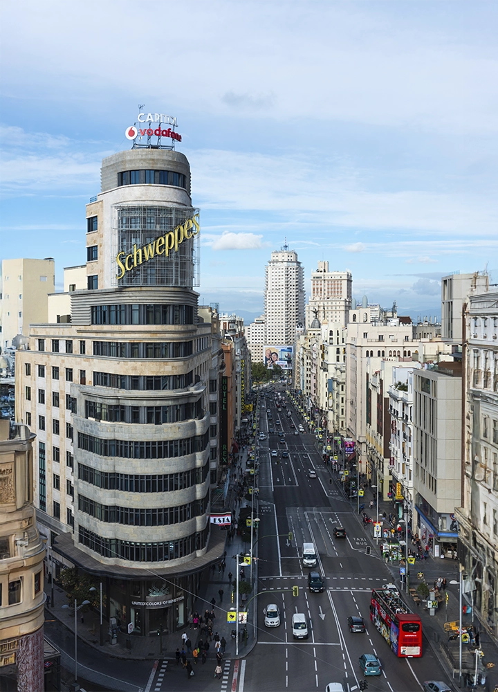 Vista desde arriba de una gran avenida urbana con mucho tráfico, flanqueada por edificios altos, destacando un edificio curvo con letreros luminosos en la parte superior a la izquierda.