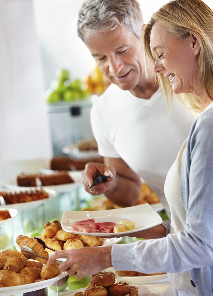 Pareja sonriente seleccionando alimentos en un buffet de desayuno, con platos de croissants, embutidos y fruta fresca en el fondo.