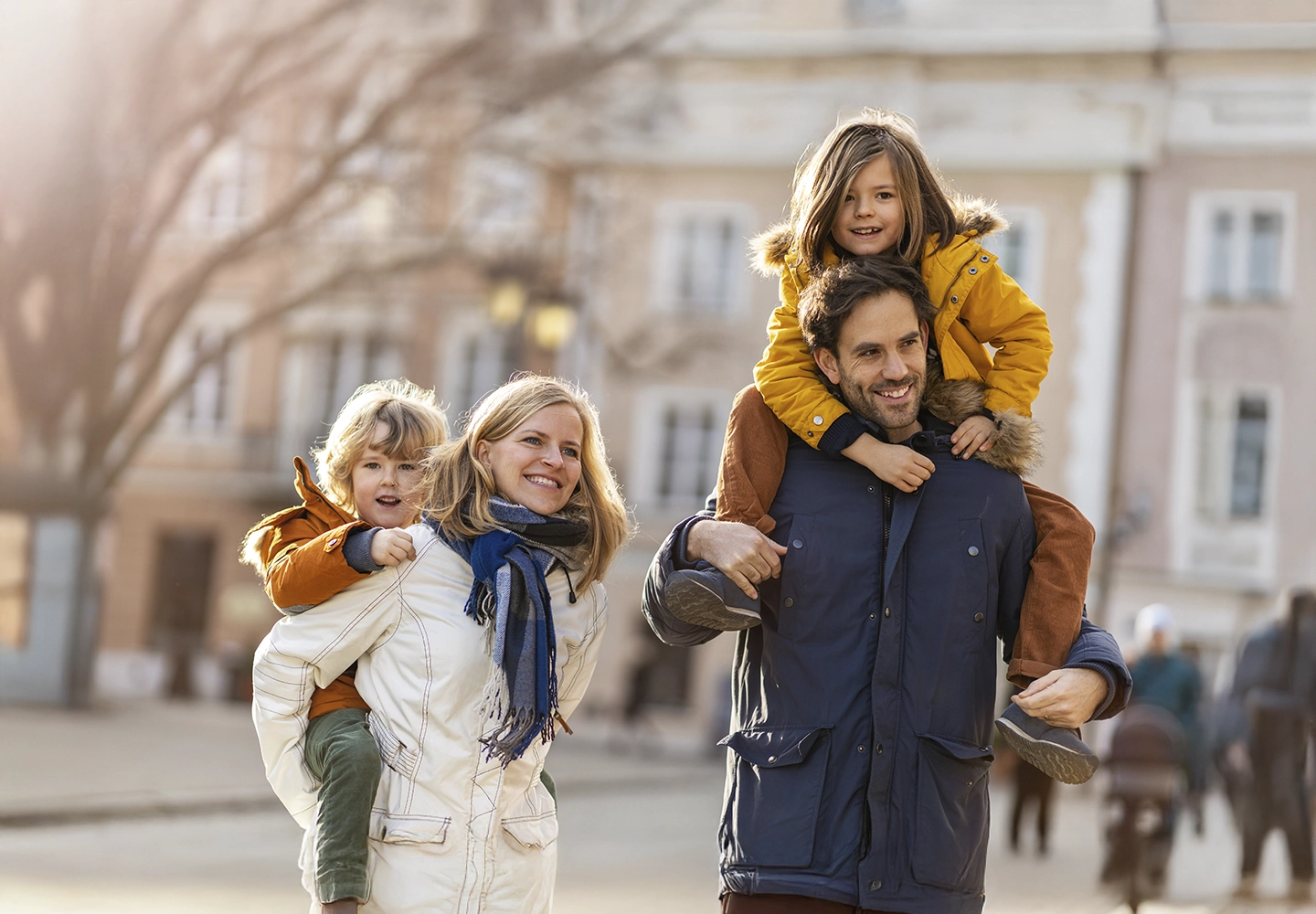 La imagen muestra a una familia disfrutando de un paseo al aire libre. Los padres llevan a sus hijos pequeños a cuestas, todos vestidos con ropa de abrigo. Están sonriendo, lo que transmite un momento de alegría y unión familiar en un entorno urbano.
