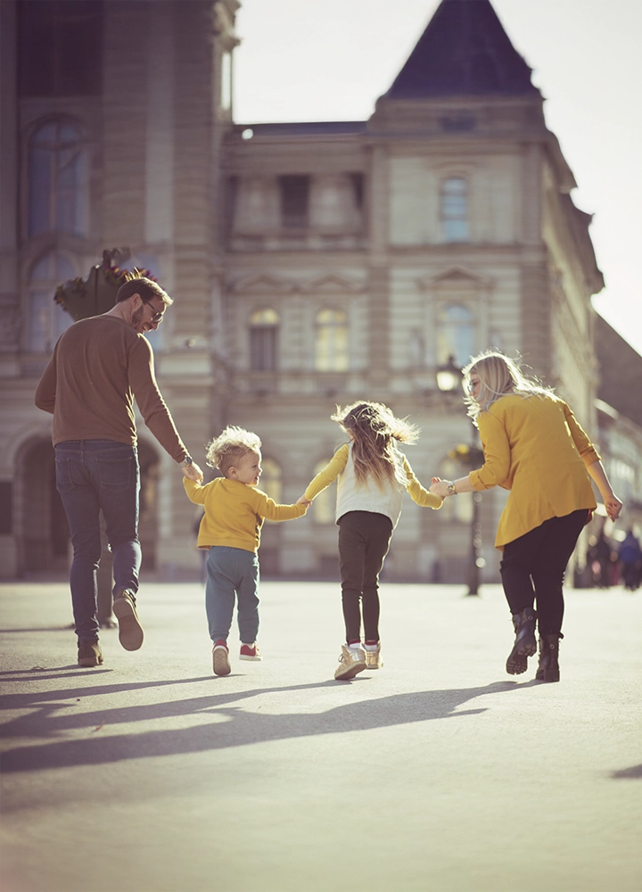 Una familia de cuatro personas , dos adultos y dos niños, camina alegremente de la mano en una plaza urbana con arquitectura clásica al fondo. Todos visten ropa otoñal en tonos cálidos, destacando el amarillo, que crea una sensación de unidad y armonía. La imagen transmite alegría, unión familiar y libertad, con una atmósfera luminosa y un entorno urbano elegante, posiblemente en una ciudad europea.