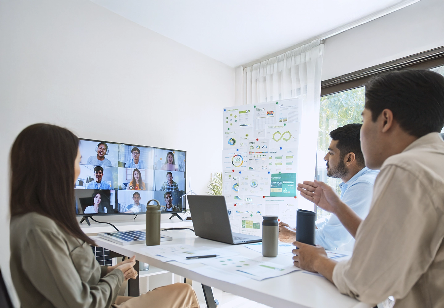 Reunión de trabajo en una oficina moderna. Tres personas participan de manera presencial, mientras en la pantalla grande se muestran los rostros de varios compañeros conectados en videollamada. En la mesa hay portátiles, termos y documentos gráficos sobre sostenibilidad. La sala está iluminada con luz natural gracias a una gran ventana.