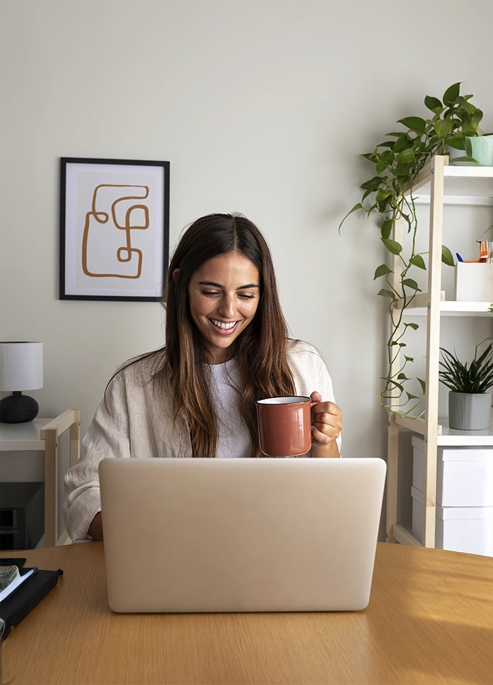 Mujer joven sonriente trabajando desde casa con un ordenador portátil, sosteniendo una taza de café. La habitación está decorada con plantas y estanterías minimalistas.