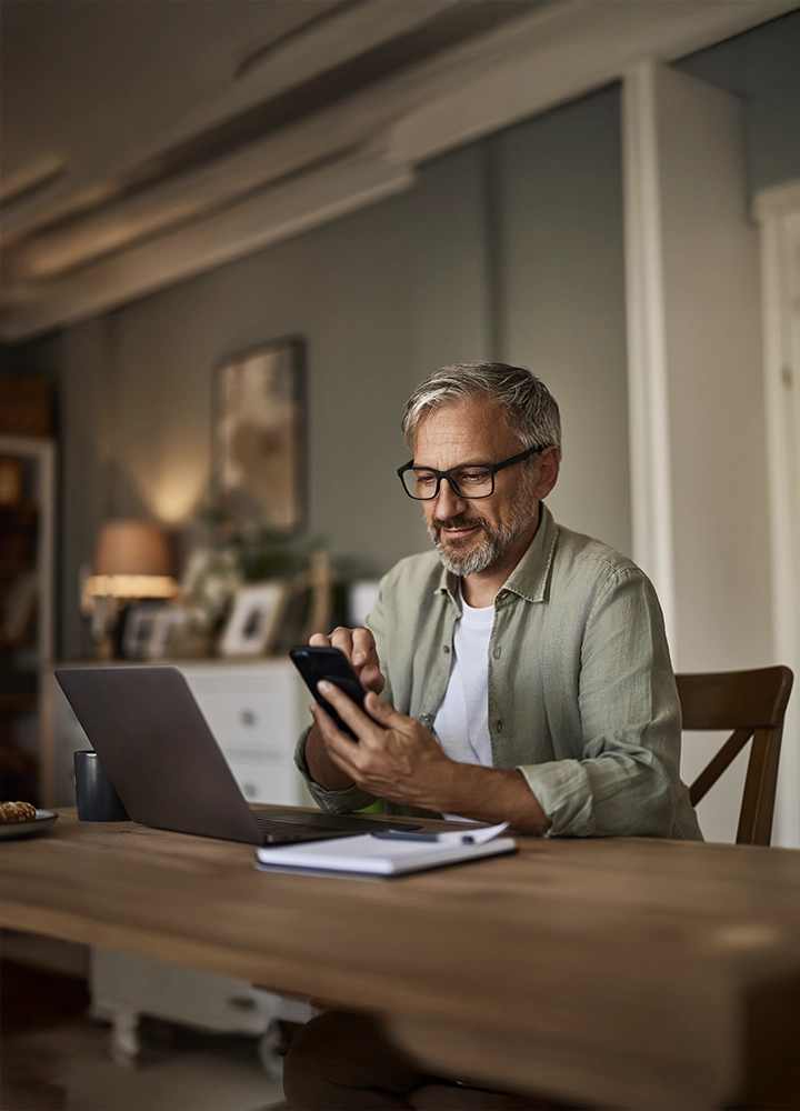 Hombre con gafas trabajando desde casa, utilizando su teléfono móvil mientras revisa su portátil. El entorno es acogedor y está decorado con muebles de madera, lámparas cálidas y fotografías familiares.