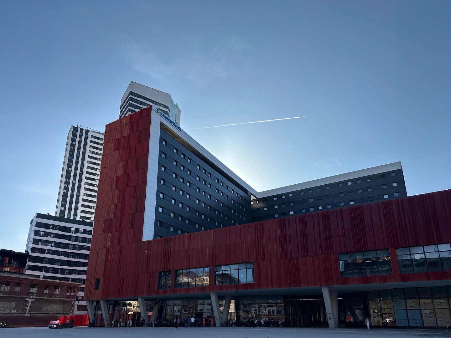 Hotel ILUNION San Mamés visto desde un ángulo bajo, mostrando su estructura con secciones de color rojo óxido y gris oscuro, y el logo de la cadena hotelera en la parte superior contra un cielo azul claro.