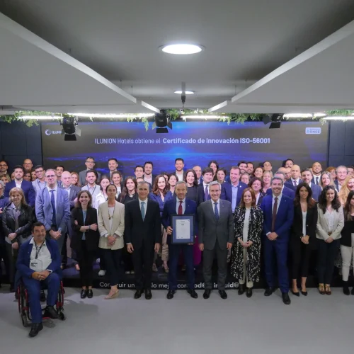 Grupo numeroso de personas posando formalmente frente a un cartel que indica que ILUNION Hotels ha obtenido el Certificado de Innovación ISO-56001. Al frente, varios representantes sostienen el certificado, destacando el logro institucional en un ambiente profesional.