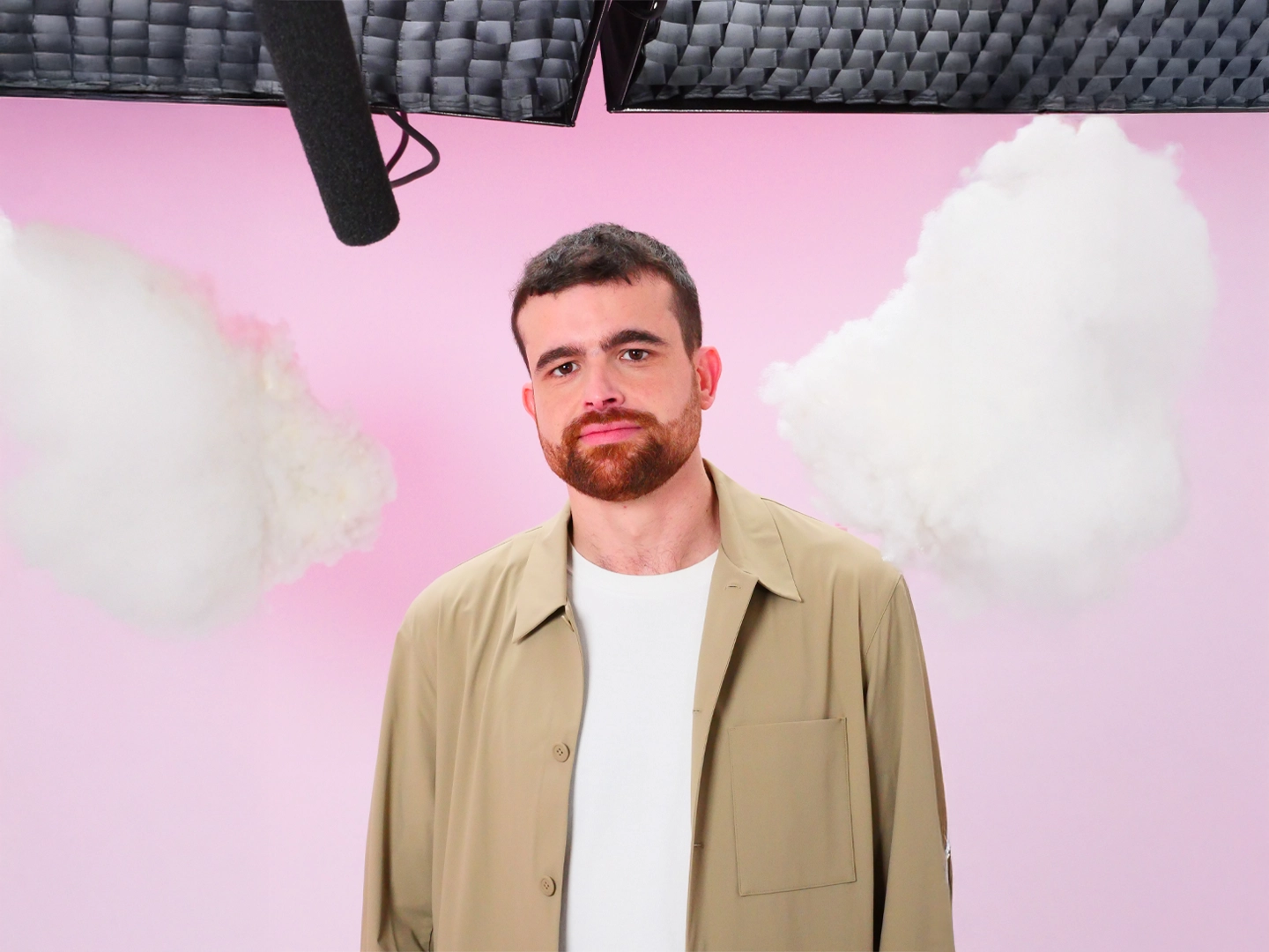 Hombre con barba corta y chaqueta beige sobre una camiseta blanca, mirando a la cámara. El fondo es un cielo rosado con nubes blancas. En la parte superior izquierda, se ve un micrófono de estudio negro.