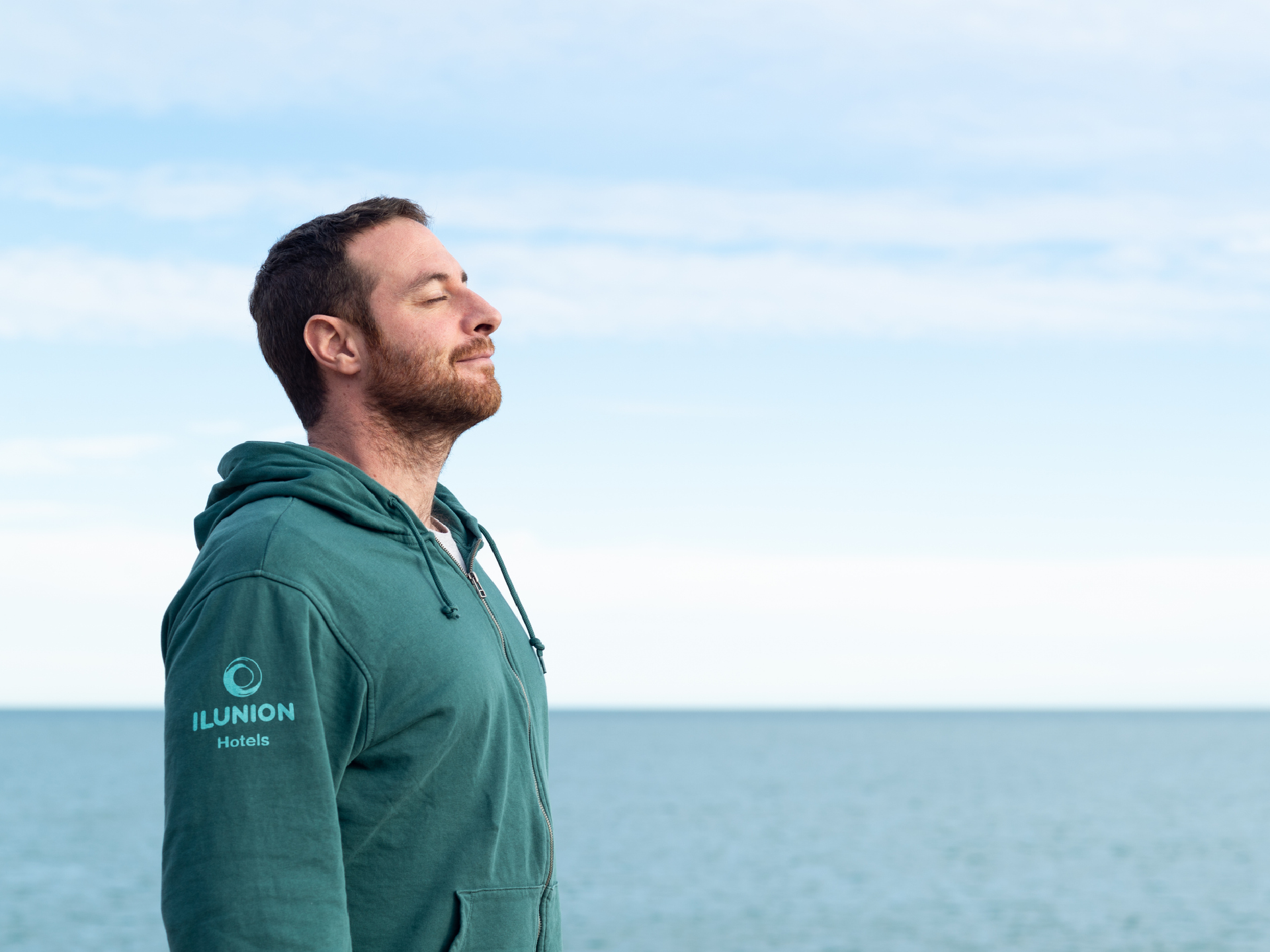 Fotografía de un hombre con barba y una sudadera verde azulado con el logo "ILUNION Hotels" en la manga, de pie con los ojos cerrados frente a un mar tranquilo y un cielo nublado. El hombre parece estar respirando profundamente o disfrutando del aire fresco.