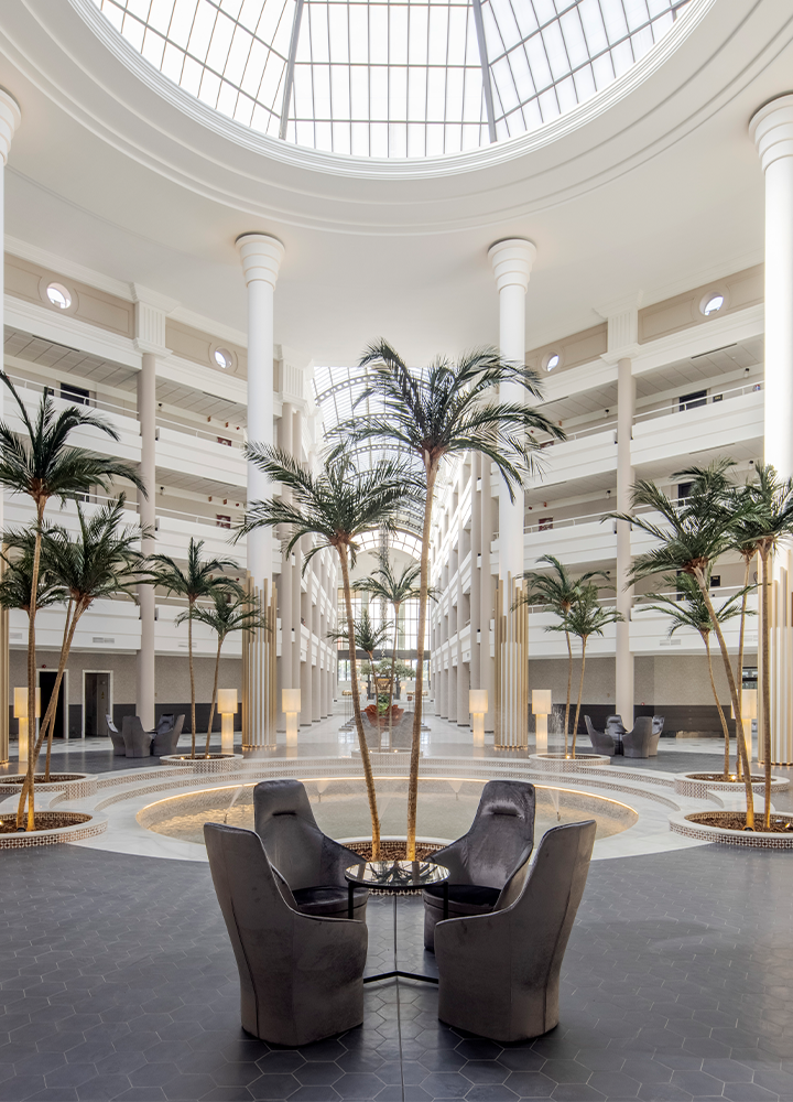 Patio interior cubierto por una cúpula de cristal, con columnas blancas, palmeras decorativas y una fuente en el centro rodeada de sillas.