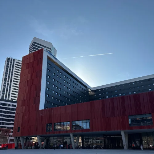 ILUNION San Mamés Hotel seen from a low angle, showcasing its structure with sections in rust-red and dark gray, and the hotel chain's logo at the top against a clear blue sky.