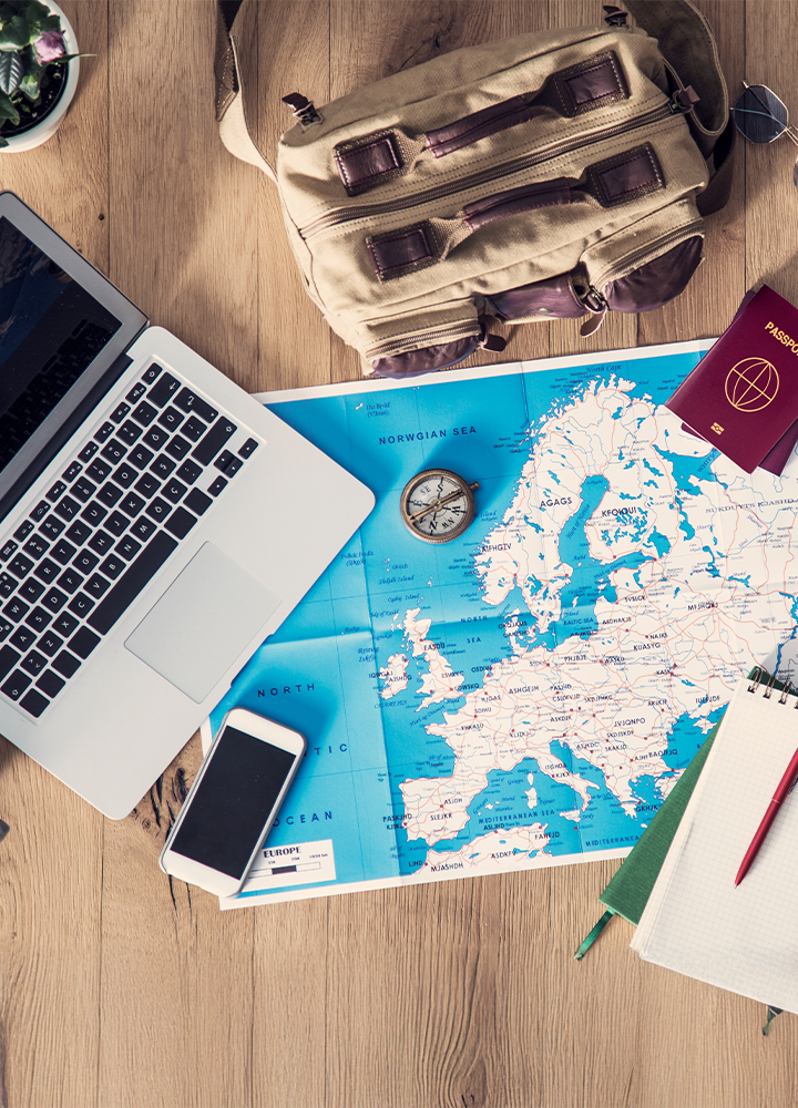 Mesa de madera con objetos como un portátil, un teléfono, un mapa, una brújula, un pasaporte y una mochila.