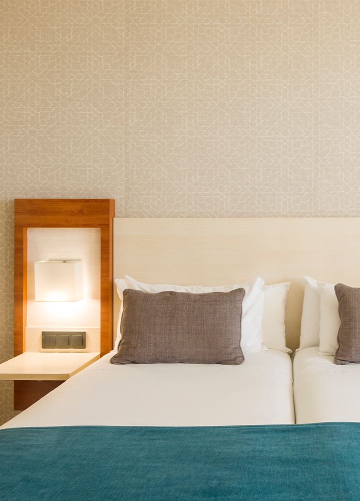 Habitación de hotel con dos camas individuales colocadas juntas. Las camas tienen sábanas blancas y están cubiertas con una manta azul en la parte inferior.
