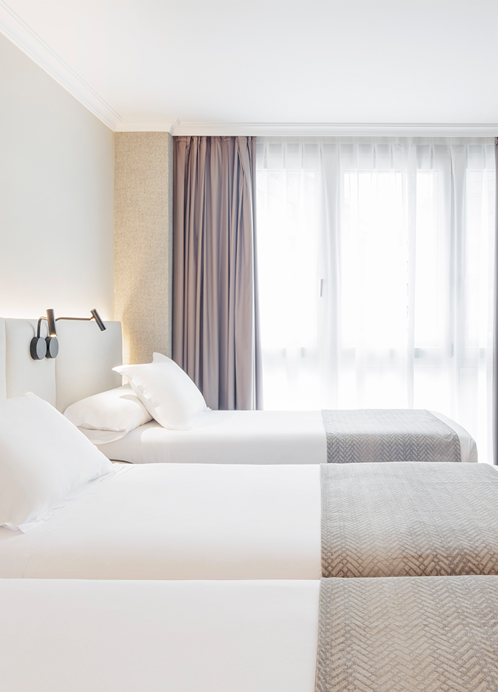Habitación de hotel con tres camas individuales, con sábanas blancas y tienen una manta gris en la parte inferior.