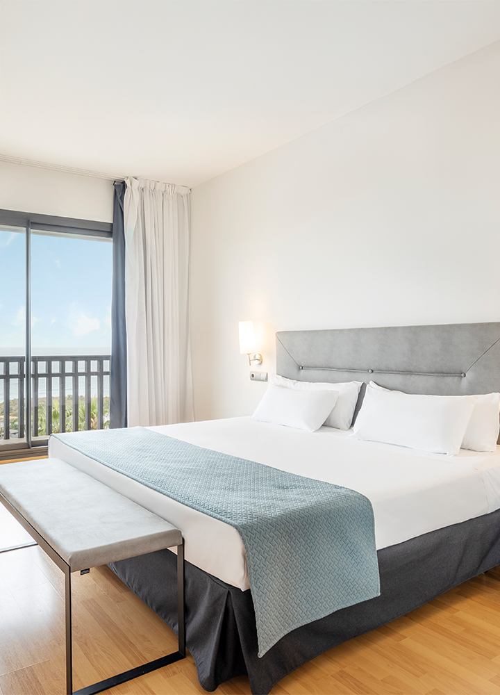 Una habitación luminosa con cama grande, lámpara encendida y ventana abierta hacia un balcón.