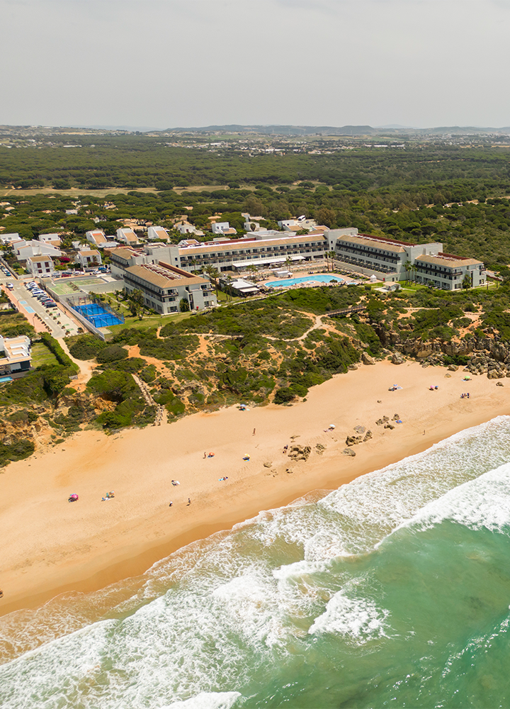 Hotel grande junto al mar, con una piscina, canchas deportivas y personas disfrutando en la playa bajo sombrillas.