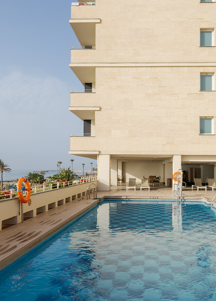 Piscina al aire libre junto a un edificio, rodeada de palmeras y con vista al mar.