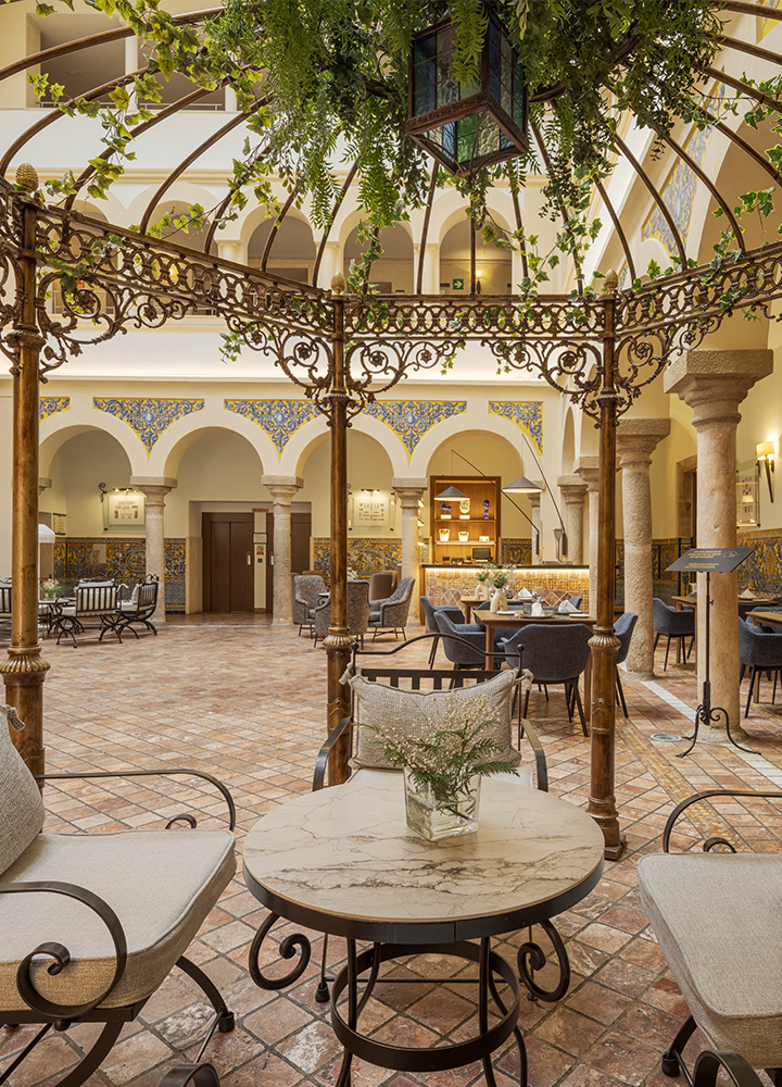 Patio interior con una arquitectura elegante y detalles decorativos. Hay una estructura de hierro forjado con detalles ornamentales, de la cual cuelga una lámpara y algunas plantas.