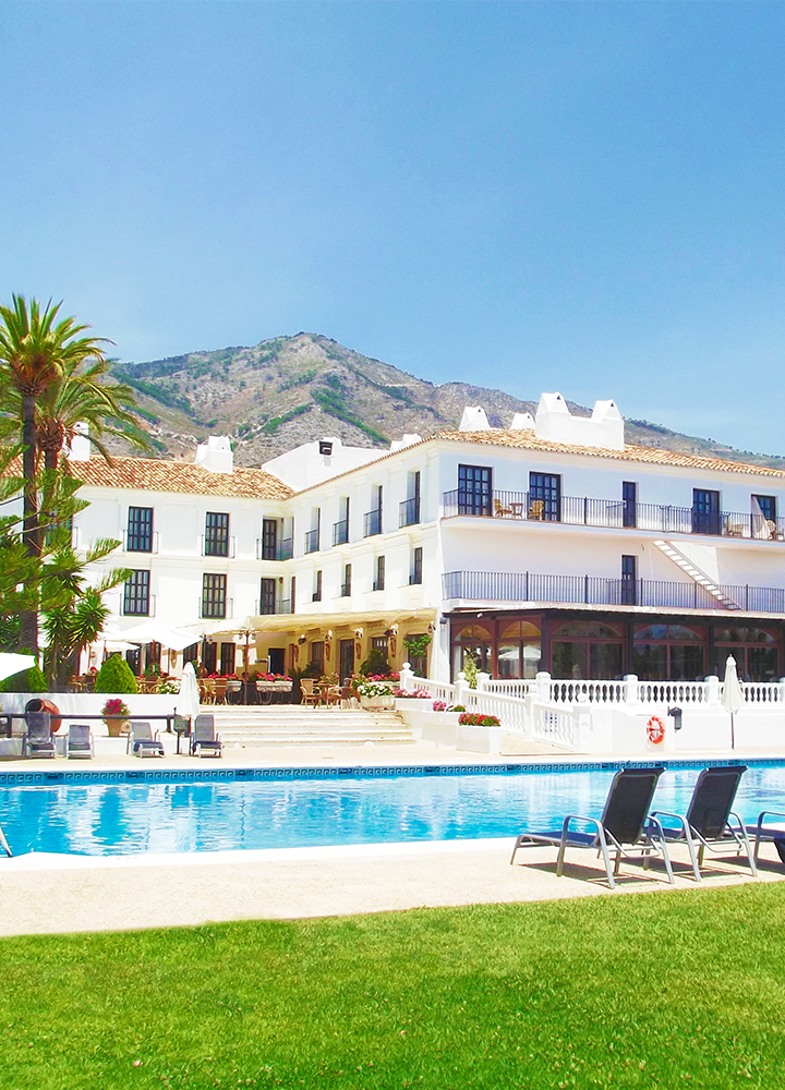 Hotel blanco con balcones detrás de una piscina azul, rodeada de mesas, sillas y una montaña al fondo.