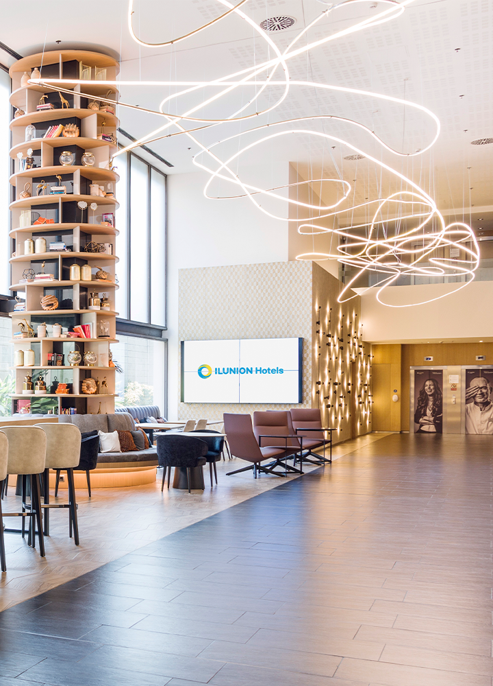 Recepción de hotel con una gran estantería circular decorativa, lámparas colgantes y una pantalla que dice "ILUNION Hotels".
