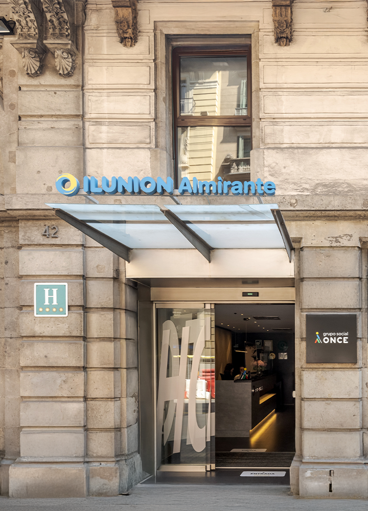 Entrada de un hotel con un toldo de vidrio, un cartel azul y amarillo que dice "ILUNION Almirante".