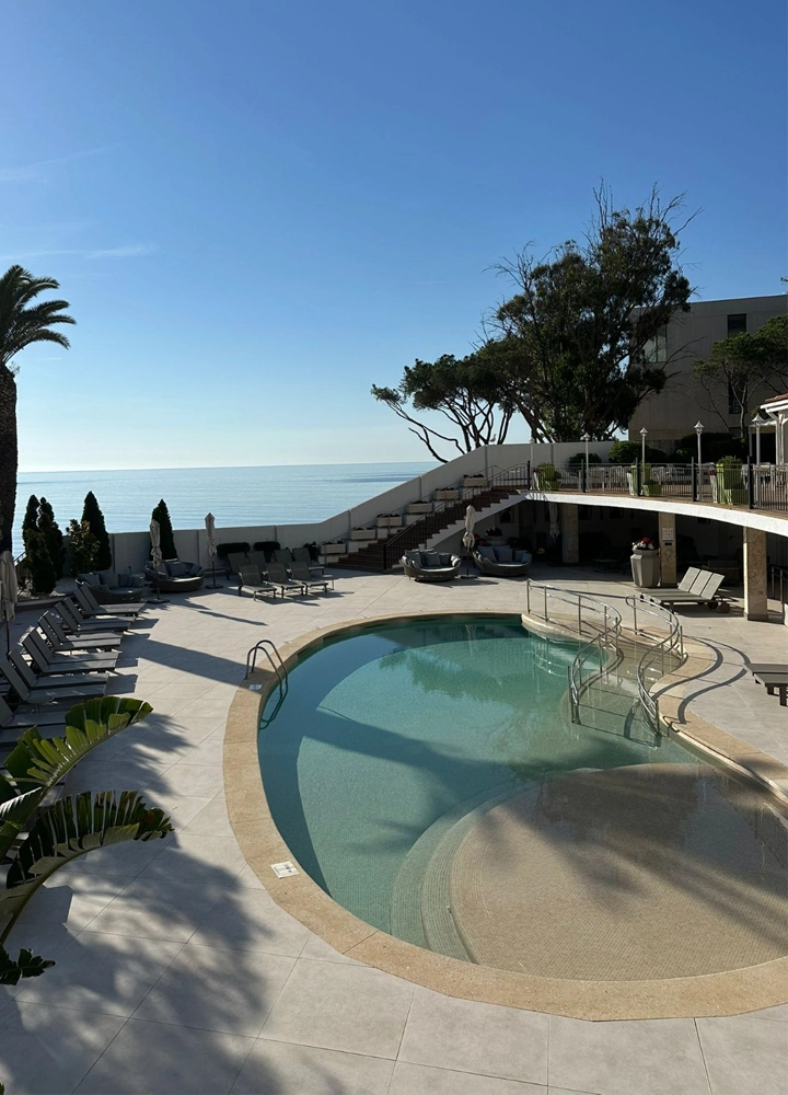 A panoramic view of an outdoor pool with the sea in the background. The pool has an irregular, tiered shape, with light blue water.