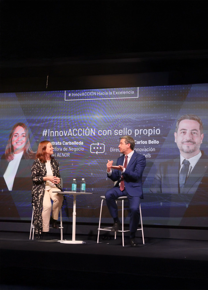 A woman and a man are seated on stools on a stage, participating in a panel discussion titled 