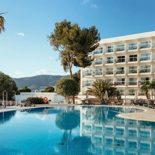 Hotel con piscina grande y luminosa rodeada de tumbonas, sombrillas y árboles.