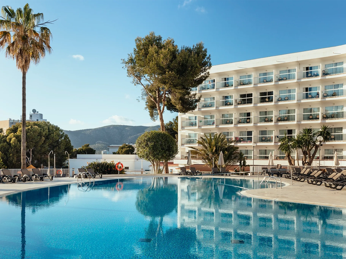 Hotel con piscina grande y luminosa rodeada de tumbonas, sombrillas y árboles.