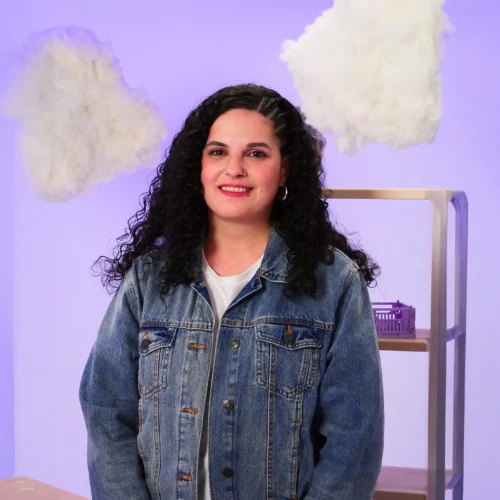 Mujer con cabello rizado y largo, vestida con una chaqueta vaquera azul y una camiseta blanca. El fondo es de un suave color morado con algunas nubes blancas dispersas, evocando quizás un ambiente onírico o introspectivo.