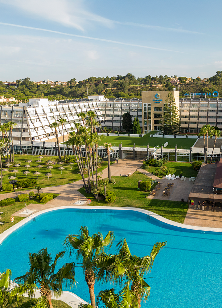 Hotel grande con piscina amplia, palmeras y jardines.