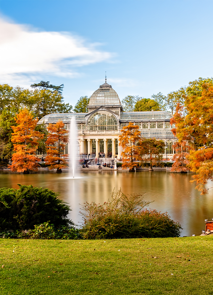 Palacio de cristal en el Parque del Retiro se refleja en un lago rodeado de árboles con hojas otoñales.