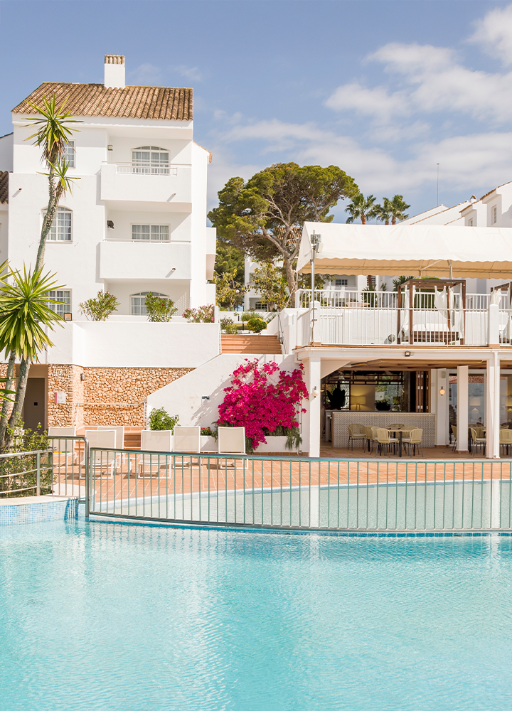 Hotel con una piscina tranquila rodeado de arbustos verdes y una buganvilla fucsia en flor.