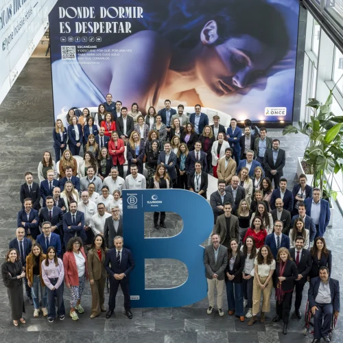 A high-angle view shows a large group of men and women of various ages posing for a photo inside a modern space with gray tiled flooring. At the lower center, a large blue ILUNION logo with a stylized 