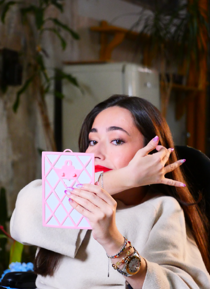 A person holding a pink object with a diamond pattern and a small lock design in the center. The individual is wearing a beige sweater and several bracelets on their left wrist, including a watch. In the background, indoor plants and furniture create a warm and decorative atmosphere. The object in the person's hands stands out for its distinctive design, particularly its texture and color. The accessories add interesting details to the composition of the image.