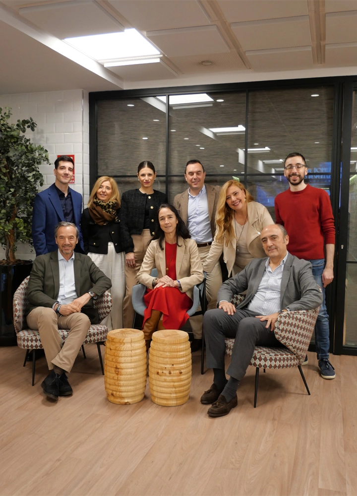 Group of nine people posing together inside a modern room with corporate decor. Three people are seated on chairs in front of two barrel-shaped designer tables, while the other six stand behind them. The room features light wooden flooring, a wall with large windows, and decorative plants on the left side.