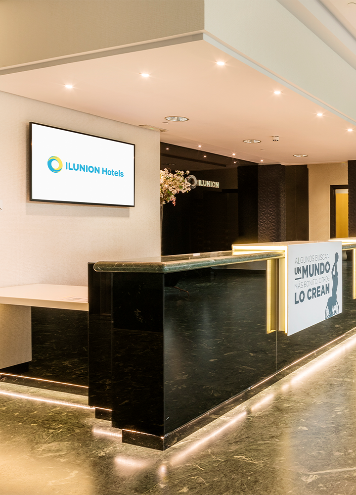 Recepción elegante de hotel con un mostrador negro brillante e iluminado. Encima, una pantalla que pone "ILUNION Hotels".