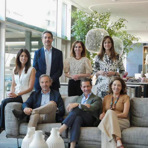 Siete personas posan en grupo dentro de un salón moderno, con plantas decorativas y lámparas colgantes; algunas están sentadas en un sofá y otras de pie detrás.