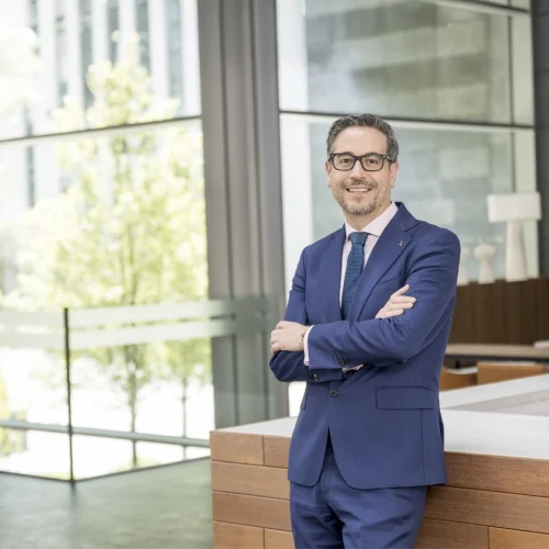 A man wearing glasses and a blue suit, smiling with his arms crossed, leaning against a chair in a modern, bright office space with large windows.