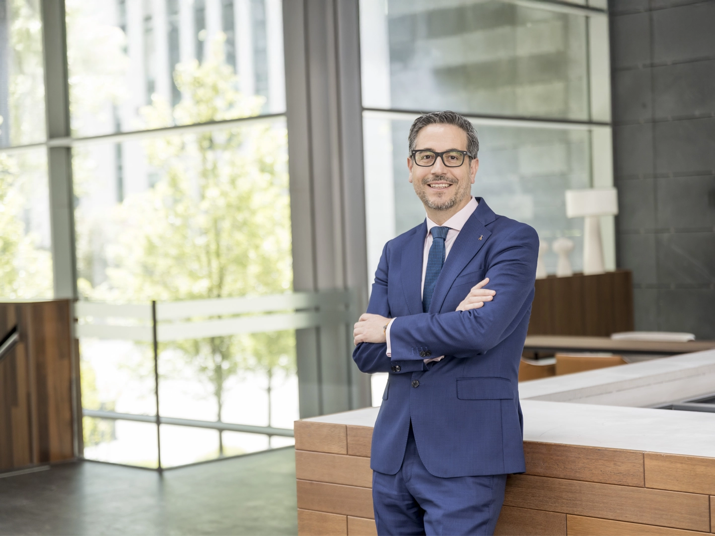 A man wearing glasses and a blue suit, smiling with his arms crossed, leaning against a chair in a modern, bright office space with large windows.