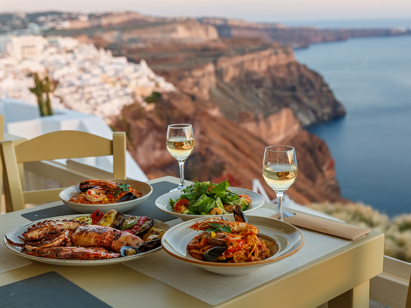 Mesa al aire libre con platos de comida y copas de vino frente al mar, con un pueblo blanco en lo alto de unos acantilados.