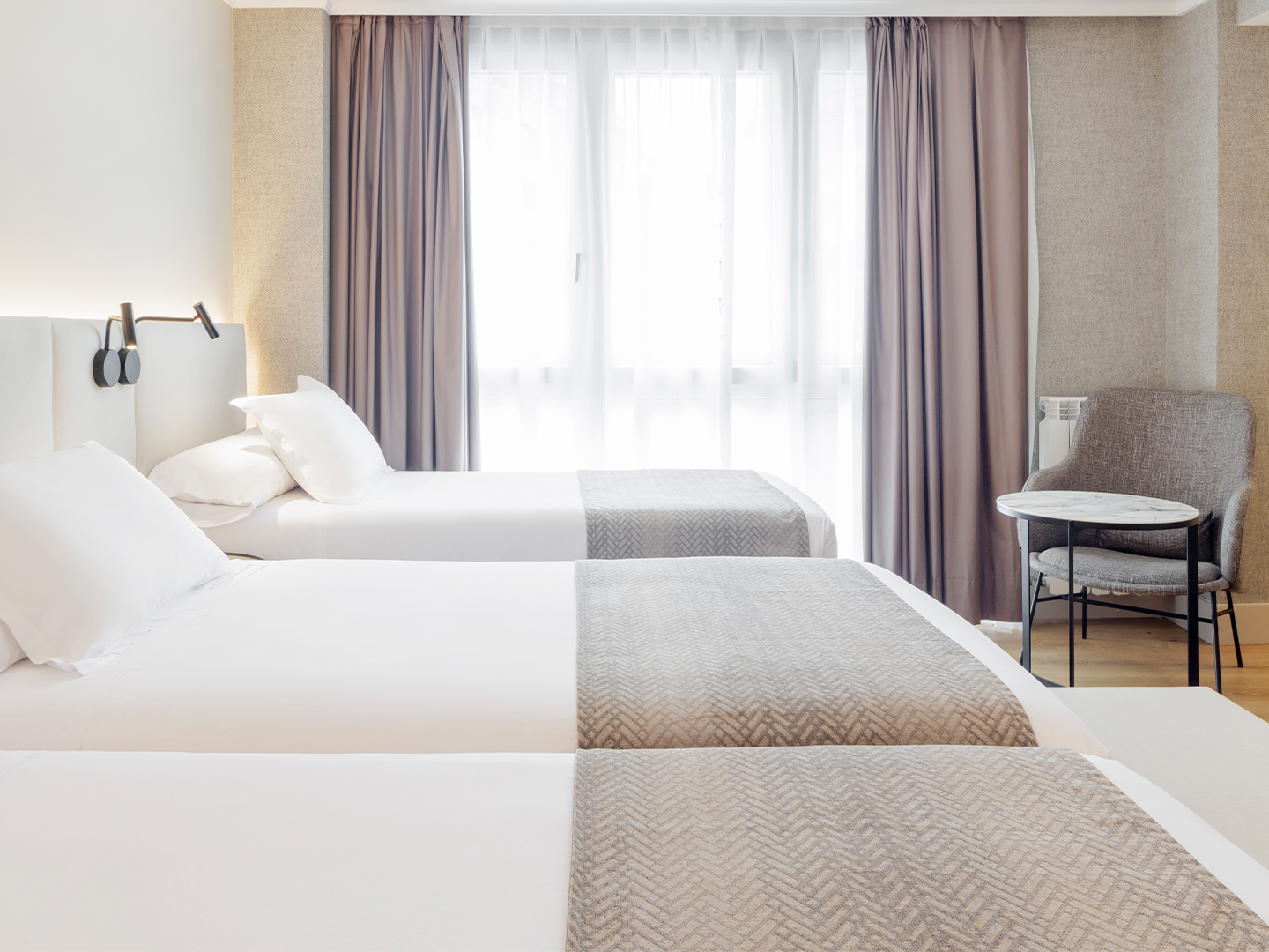 Habitación de hotel con tres camas individuales, con sábanas blancas y tienen una manta gris en la parte inferior.