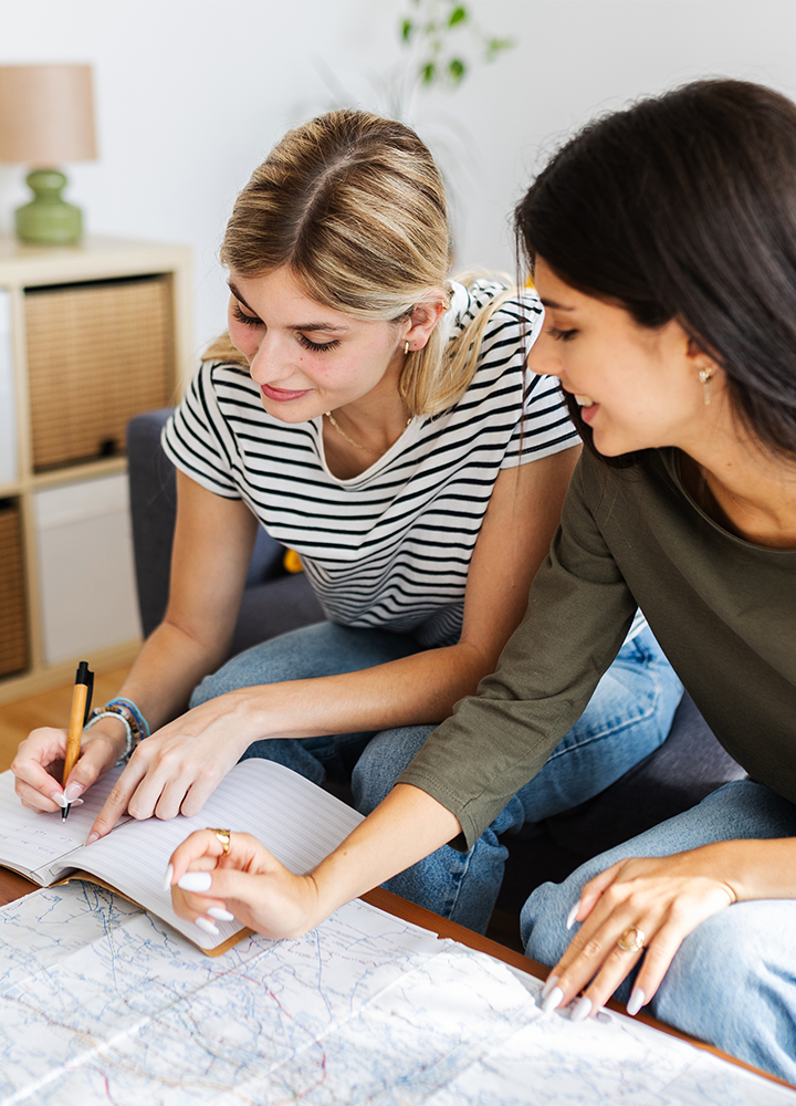 Dos personas sentadas en un sofá. Una escribe en un cuaderno y la otra señala un mapa extendido sobre una mesa.