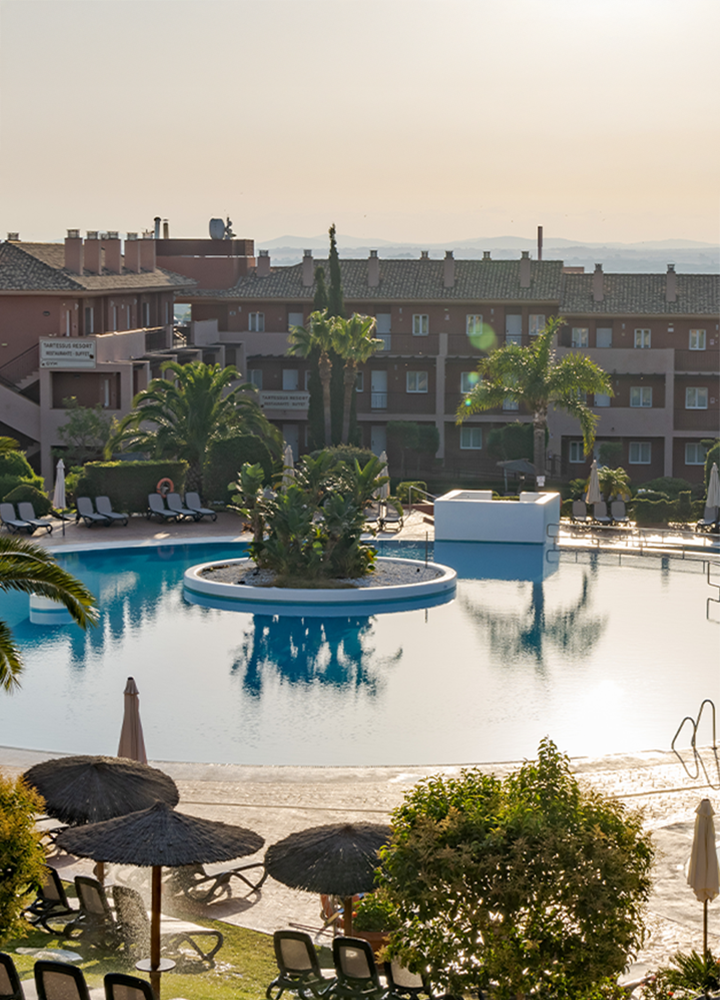 Hotel con gran piscina con forma circular que rodea un jardín central, con tumbonas y sombrillas alrededor.