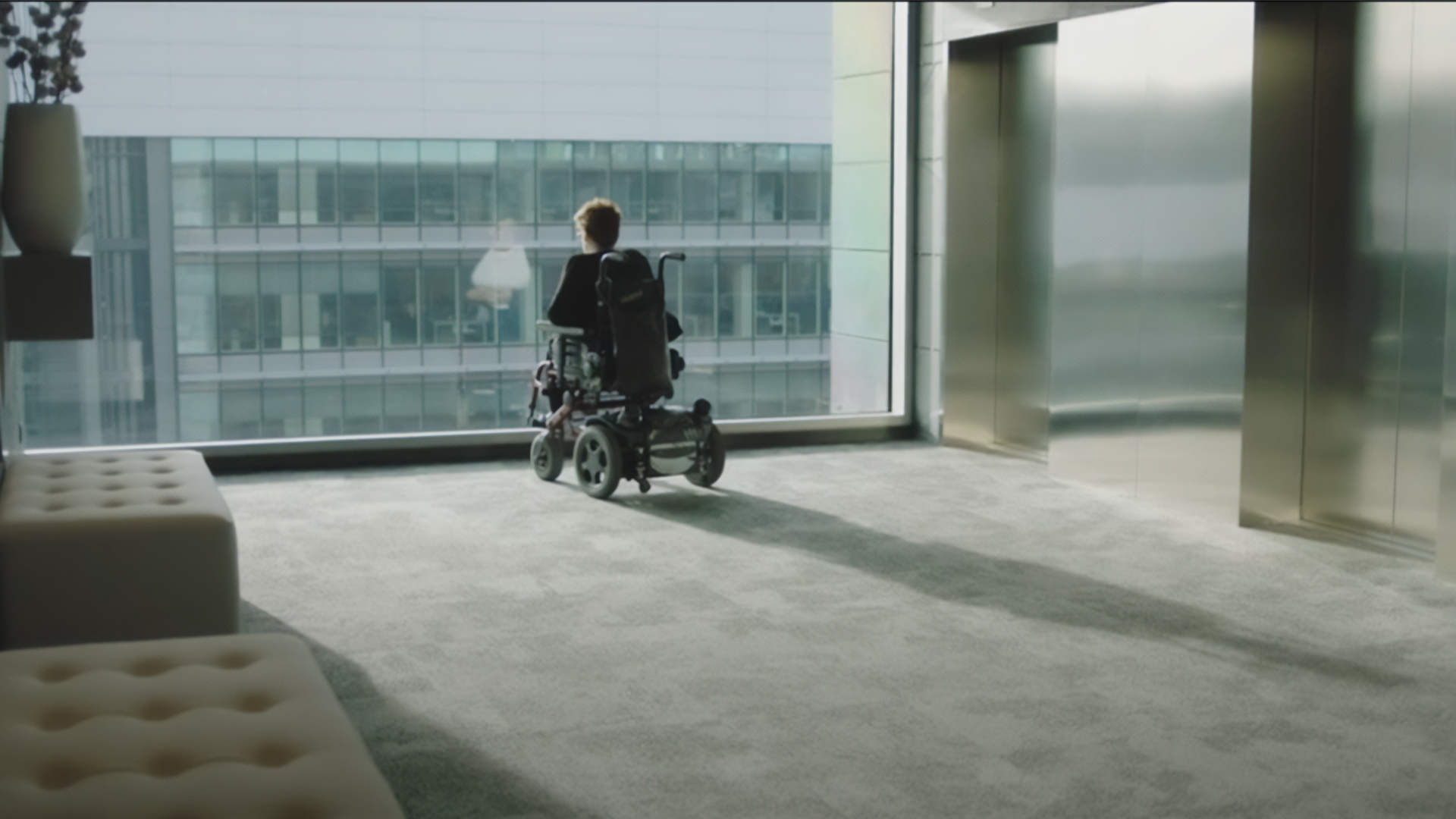 Persona en silla de ruedas viendo el exterior desde una gran ventana acristalada en un edificio moderno.