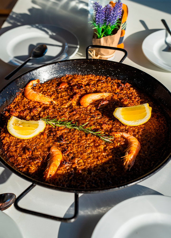 Una gran sartén de paella humeante, con arroz dorado, gambas grandes, rodajas de limón y una ramita de romero en el centro.