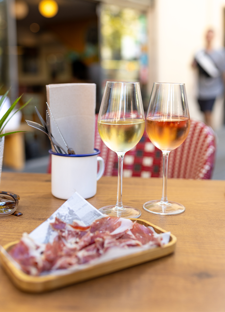 En una mesa de restaurante al aire libre hay dos copas de vino, una blanca y otra rosada, junto a un plato con lonchas de jamón.