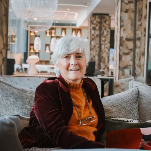 A woman with short white hair and a kind expression smiles while sitting on a sofa surrounded by cushions. She wears an elegant burgundy jacket and mustard sweater set, with glasses hanging from a cord. Behind her, there is a blurred background that appears to be a hotel lobby.